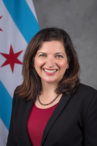 Photo of Commissioner Arfa. A smiling woman with brunette hair wearing a black blazer over a red shirt. The chicago flag is in the background.
